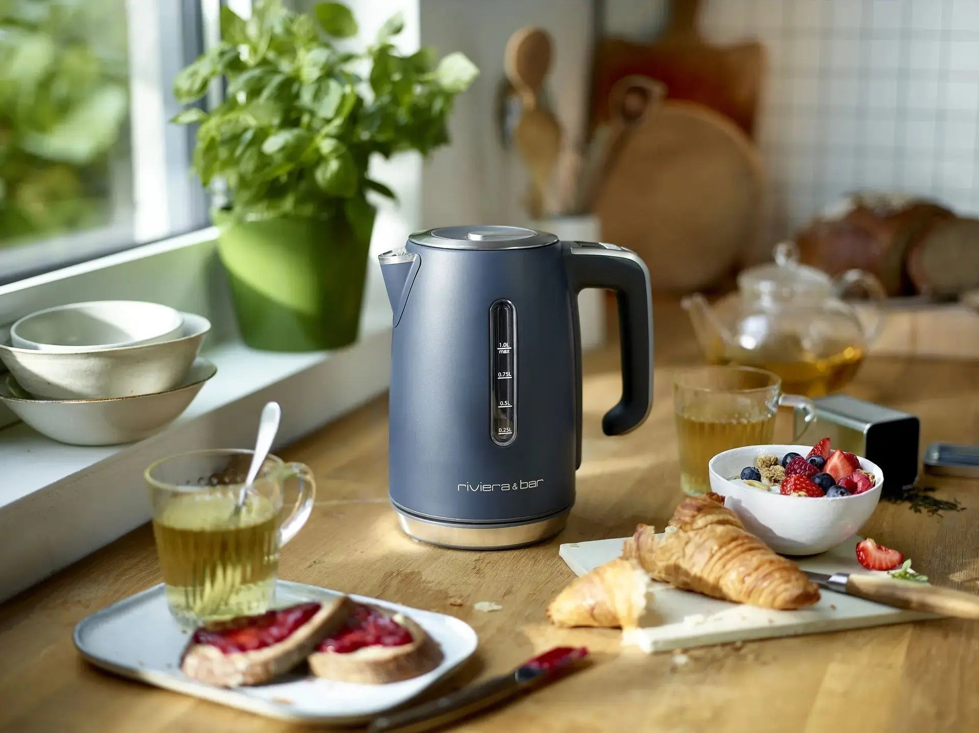 Ontbijt op een houten tafel met een grijze waterkoker, een kopje thee, toast met jam, een croissant en een kom ontbijtgranen met rode vruchten.