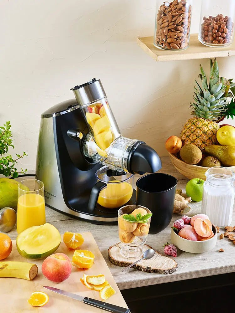 Close-up van een roestvrijstalen en zwarte sapcentrifuge, omringd door vers fruit en bakjes, op een lichthouten tafel.