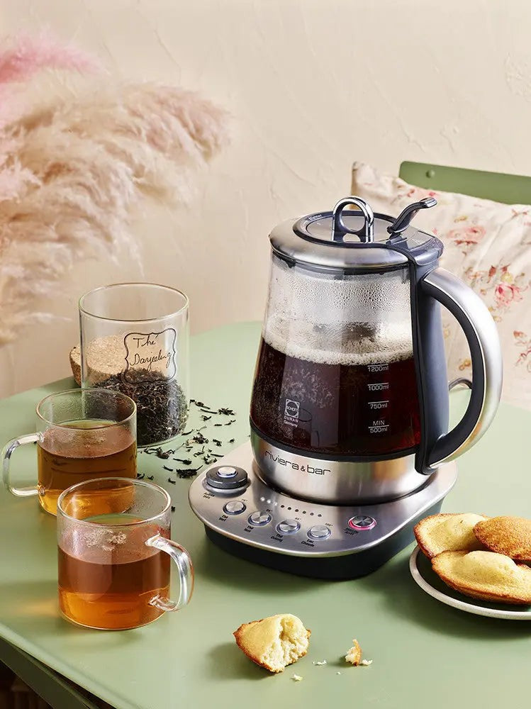 Afbeelding van een elektrische theepot van Riviera & Bar, vervaardigd uit glas en roestvrij staal, vergezeld van theekopjes, een doos Darjeeling-thee en madeleines, op een groene tafel.