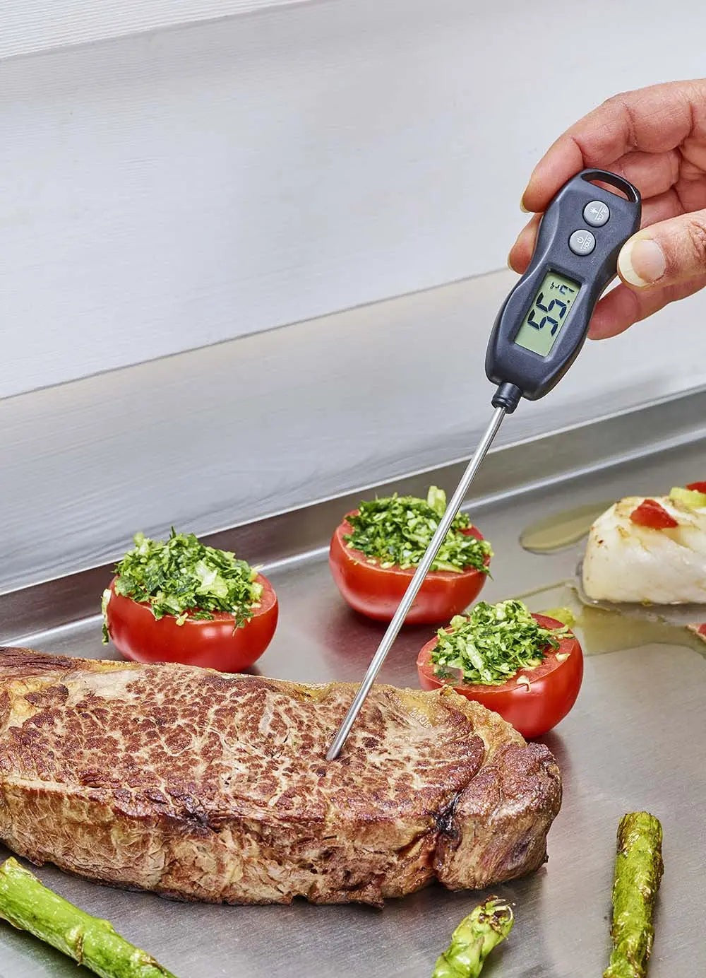 Kookthermometer voor het meten van de temperatuur van een gebakken biefstuk, gevulde tomaten en asperges in de buurt.